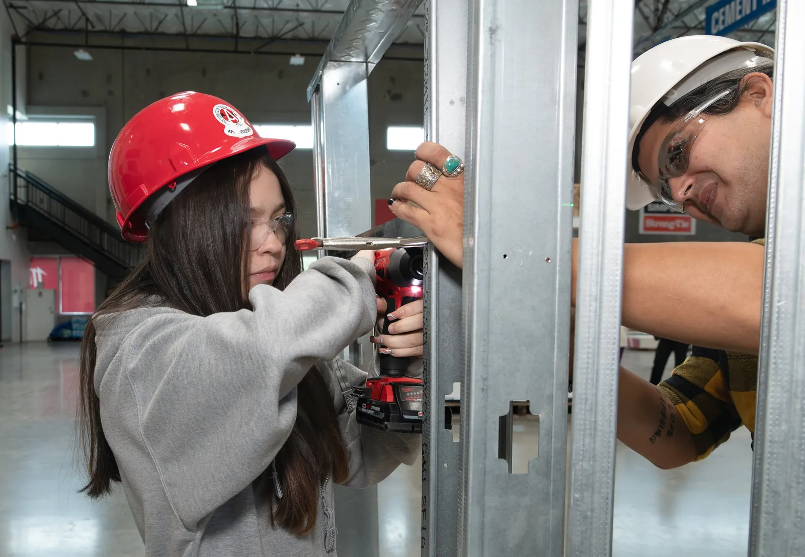 A student wearing a hard hat and safety glasses working on a metal frame with a power drill
